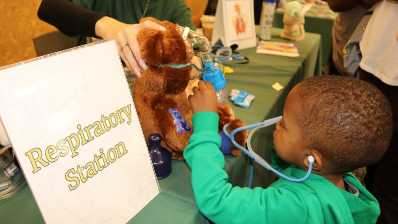 A child uses a toy stethoscope to listen to a stuffed bear's heart at a table with a "Respiratory Station" sign.