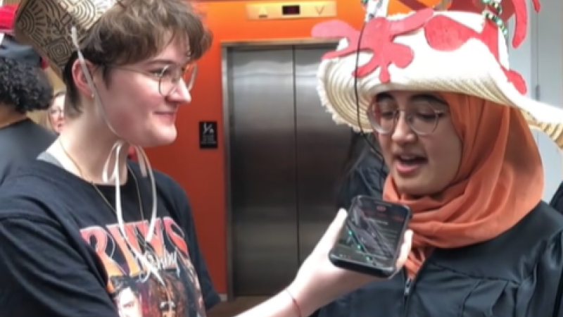 Two Smith College students wearing homemade hats. One holds a phone out to the other as a microphone.