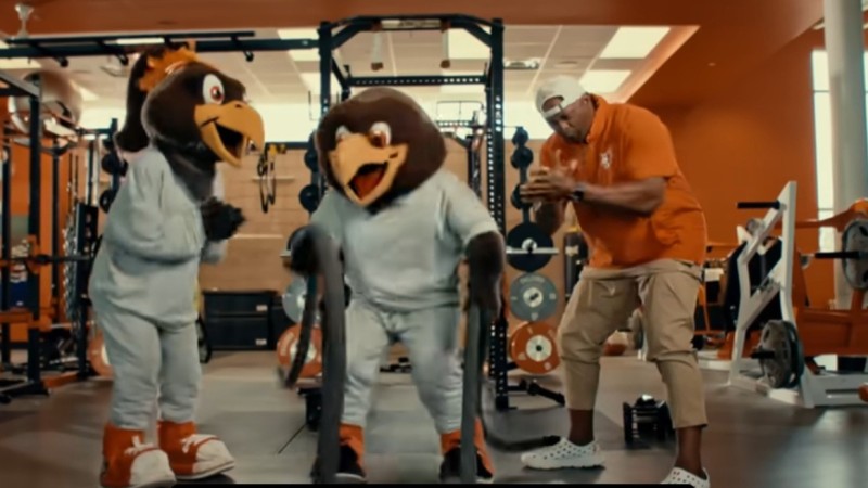 A screenshot from "Super Freddie." Two falcon mascots and a man train in a campus gym.