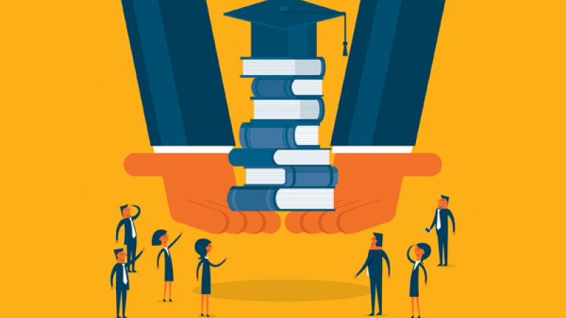 Students standing around a stack of books with a graduation cap on top