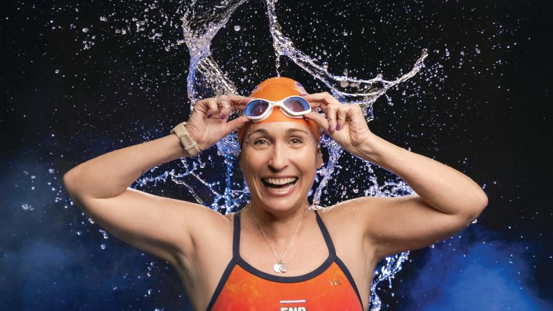 A headshot of Laurie Craigen who wears an orange swimsuit and cap, her hands raised to her goggles on her forehead.