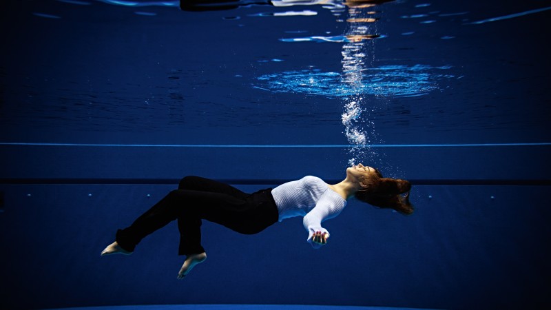 A girl dressed in business attire lays suspended and submerged in a pool. Air bubbles float from her mouth to the surface.