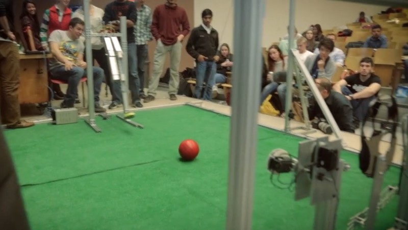 Students gather around a constructed field to watch mechanically engineered robots play with a red ball.
