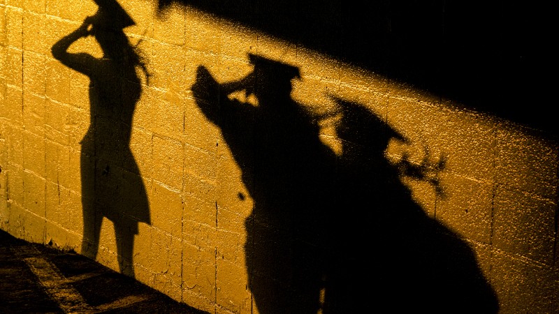 Against a brick wall are the shadows of three people in commencement dress illuminated by golden light.