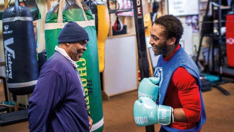 Walter Davis, wearing light blue boxing gloves, jokes with his coach Scott Brown in a boxing gym.