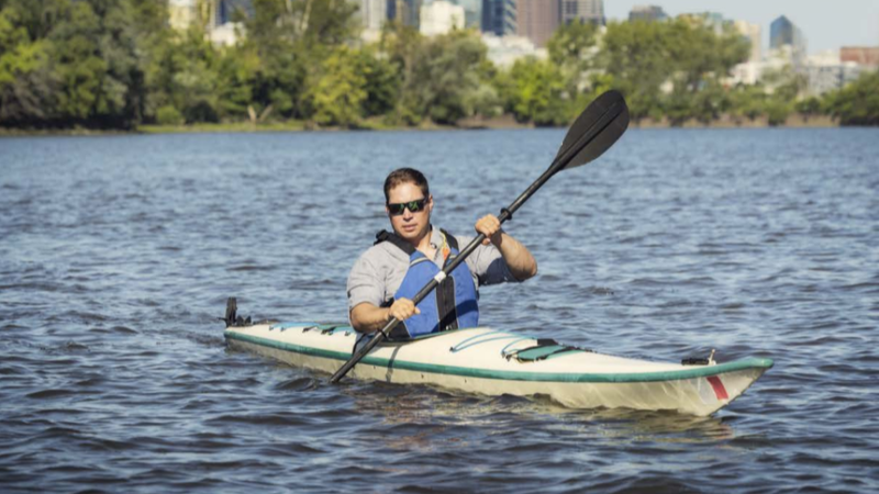 A man wearing a blue life vest kayaks in the middle of a river. In the background you can see a city skyline.