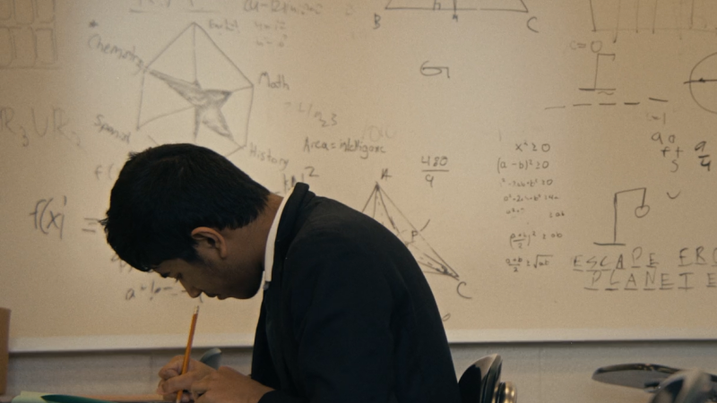 A student hunches over his desk, writing with a no. 2 pencil on a packet of papers. Behind him, a white board is covered in mathematical equations and graphs, and a game of hangman.