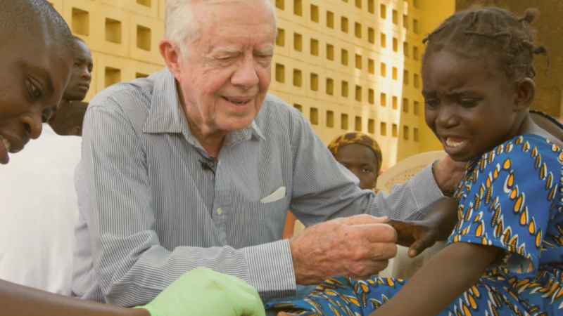 President Jimmy Carter holds the hand of a Black girl being treated for Guinea worm disease.