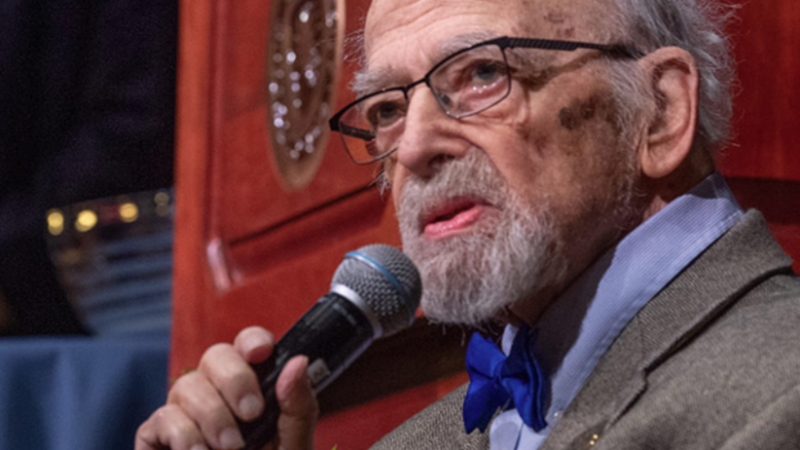 A photo of Jerry M. Lewis. He sits on a stage, holding a microphone.