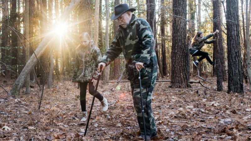 Bill Hicks uses two hiking sticks to walk on the forest's leaf-covered ground. Beside him walks a blonde girl, and another blonde girl climbs a tree truck in the background.