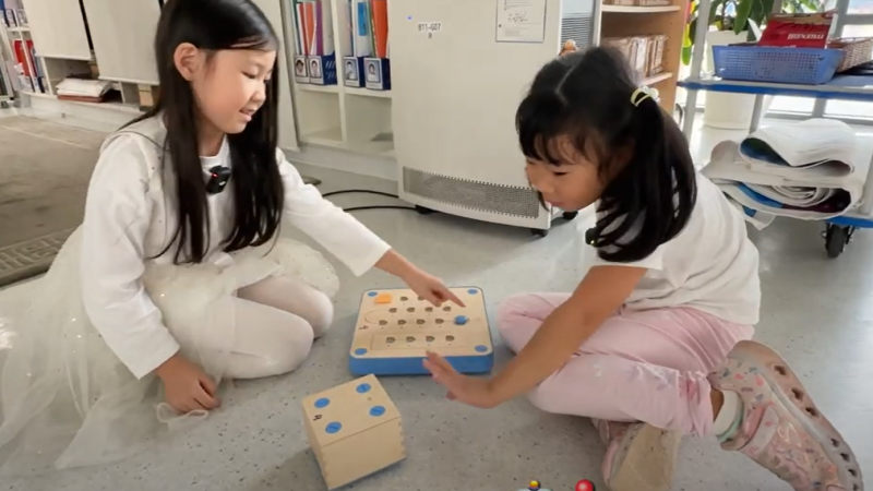 Two students in UNIS Hanoi's Early Childhood Centre sit on the ground with two block toys between them. One of them waves at the moving box, saying "Hi, Robot!"