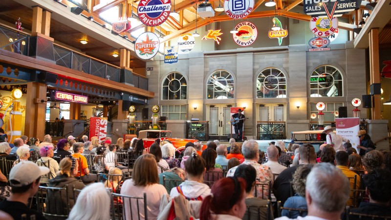 A seated audience watches a lone guitar player on stage. Vintage metal signs hang from the vaulted ceiling.