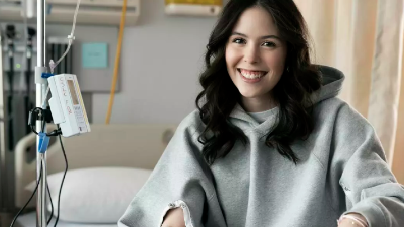 Maddison Tory smiles for a photo while sitting in a hospital bed wearing one of her IV-accessible hoodies.