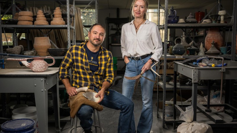 Metin Eren and Michelle Bebbers pose in a workshop filled with shelves of relics and archeology tools.