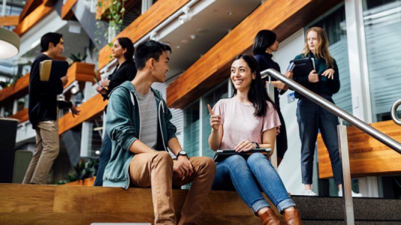 Two people sit on a staircase talking, while others move through the building behind them.