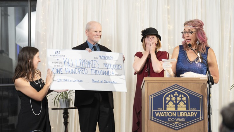 At a podium, a woman in 1920s flapper dress reads from a slip of paper. Beside her, two people hold a carboard check for $100 written out to KU Libraries and a third person covers their mouth in surprise.