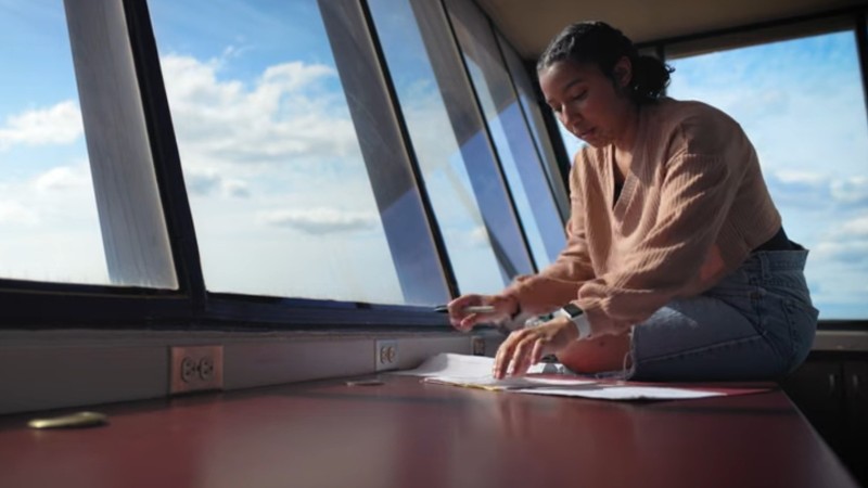 Aria Vargyas sits on a counter in a weather station sorting papers. The windows beside her show blue skies.
