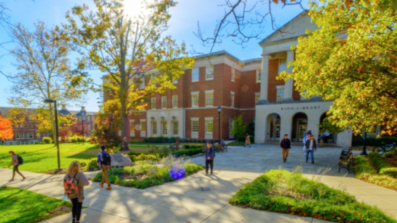 A shot of campus shows the sun rising through tree branches and students walking to and from the library.