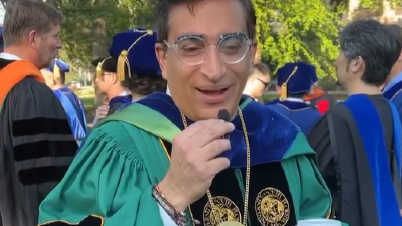 A professor dressed for commencement in green robes holds a tiny mic and answers a question for the camera.
