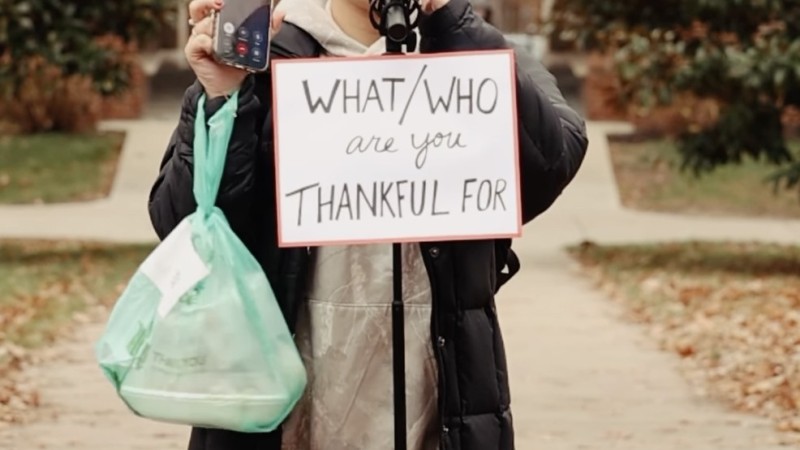 A screenshot from Miami University's "Who or What Are You Thankful For?" video shows a girl in a winter coat holding her phone up to camera as she speaks into a microphone, saying, "I'm on the phone with her right now."