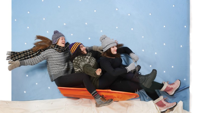 Three girls sit on a sled wearing winter hats, sweaters, and snow boots. They are posed to look like they are in motion sledding down a hill.