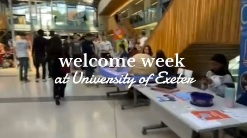 A screenshot from a video of the interior of a campus building with tables set up along its edges. The text, "welcome week at University of Exeter" has been placed at the center of the screen.